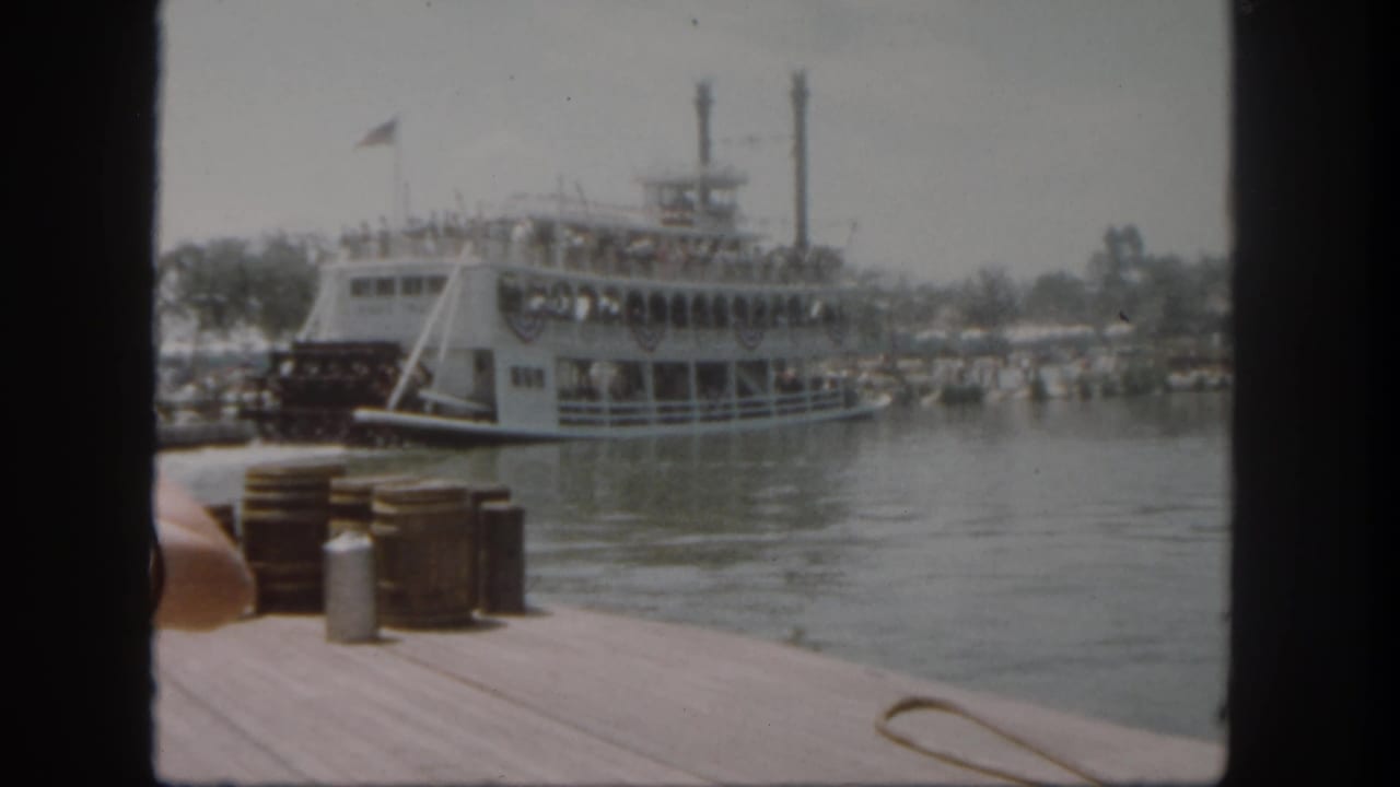 Paddlewheel boat leaving a wooden dock with barrels and rope in the foreground.