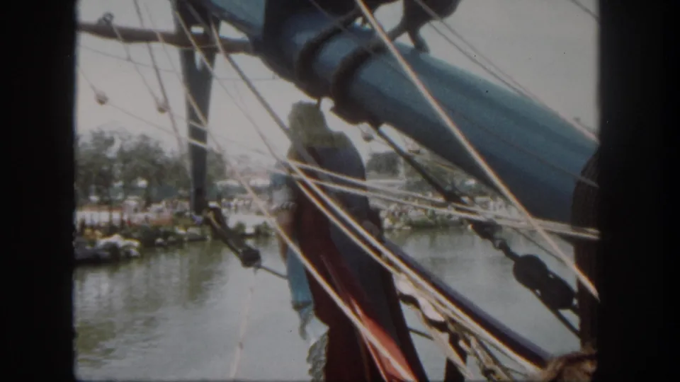 From a nearby vessel or attraction, rigging and a large boom cut across a 1962 Disneyland waterfront view with trees and water below.
