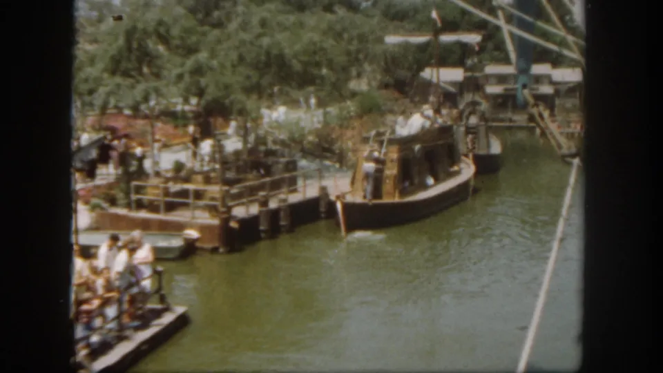1962 Disneyland California home-movie view of a small boat arriving near a dock, with guests, railings, trees, and green water visible.