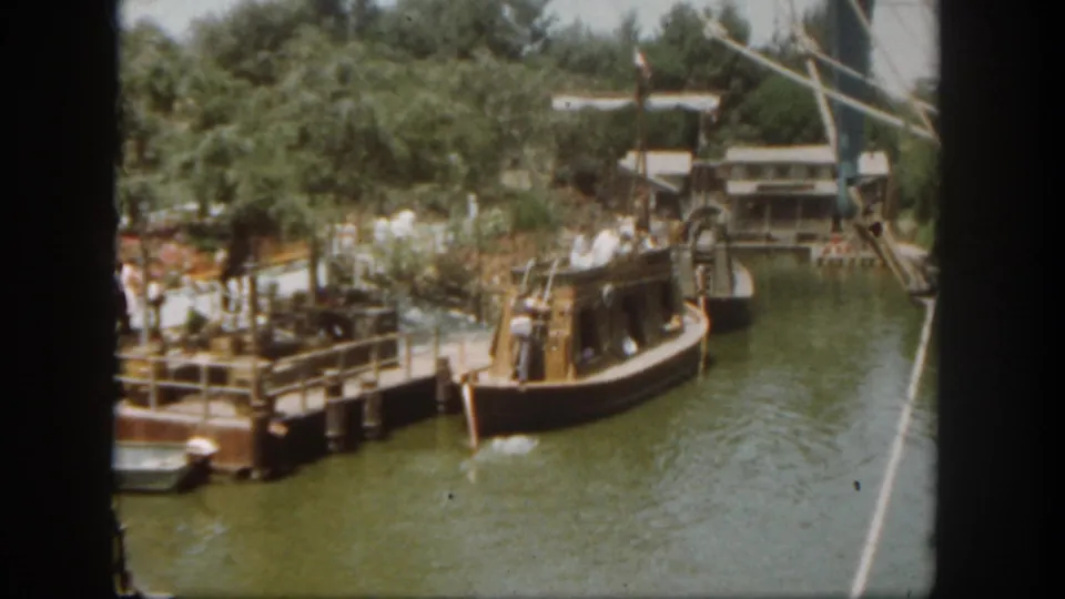 1962 Disneyland California frame showing a boat at the dock area, with visitors, railings, trees, green water, and rigging at the edge.