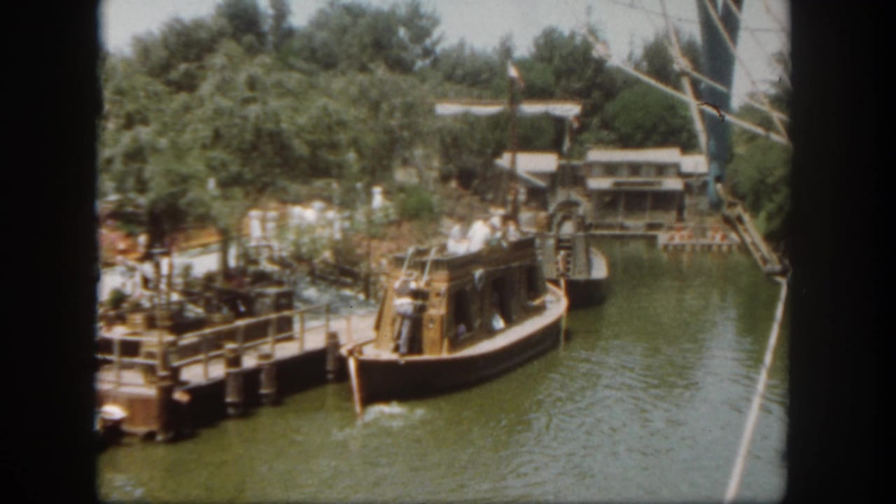 Wooden boat passes a dock on a landscaped waterway with trees and structures beyond.