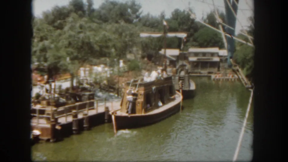 1962 Disneyland home-movie frame showing a wooden boat near a dock on a green waterway with trees and park structures in the background.