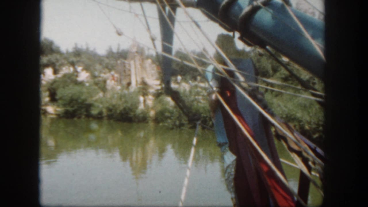 View from a boat with rigging at right, green water, trees, and structures along the bank.