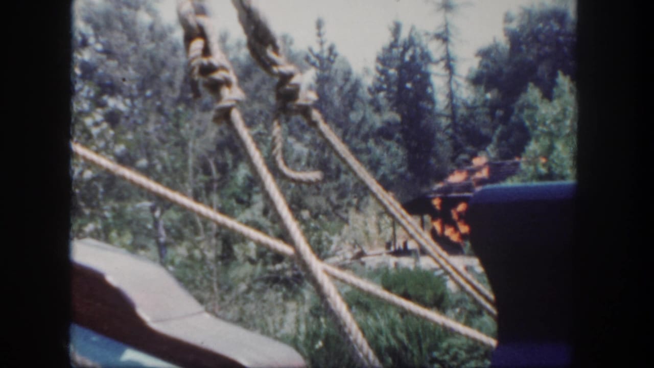 1962 Disneyland ride view showing rope supports in the foreground, wooded scenery, a partial passenger, and a staged fire effect in the distance.