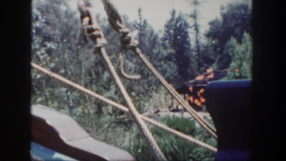 1962 Disneyland ride view showing rope supports in the foreground, wooded scenery, a partial passenger, and a staged fire effect in the distance.