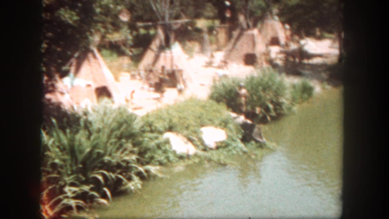 Teepees stand behind shrubs beside a green waterway in bright sun.