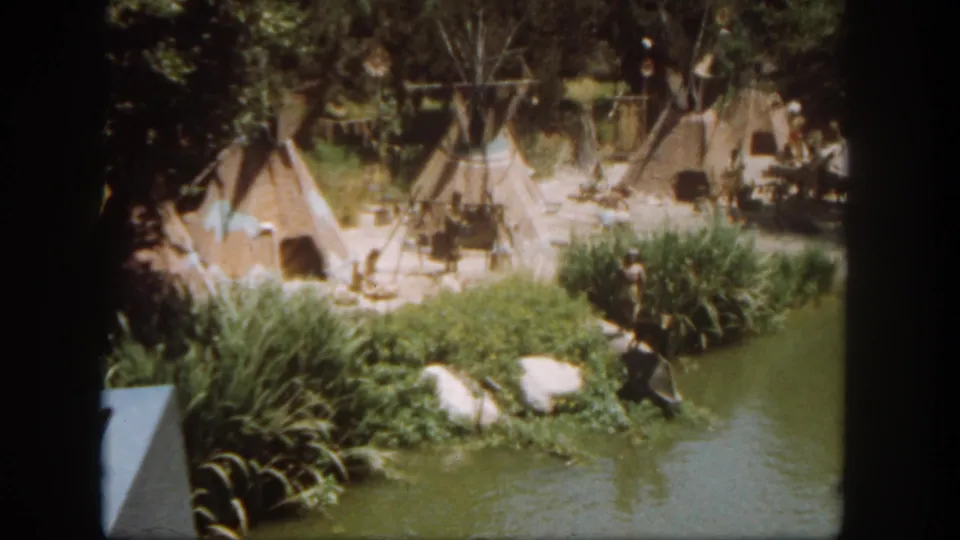 Visitor-shot 1962 Disneyland frame of a themed teepee display with poles, riverbank foliage, rocks, and calm water.