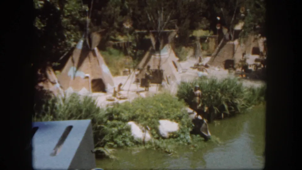 1962 Disneyland 8mm frame showing teepees, wooden poles, rocks, shrubs, green water, and a foreground structure edge.