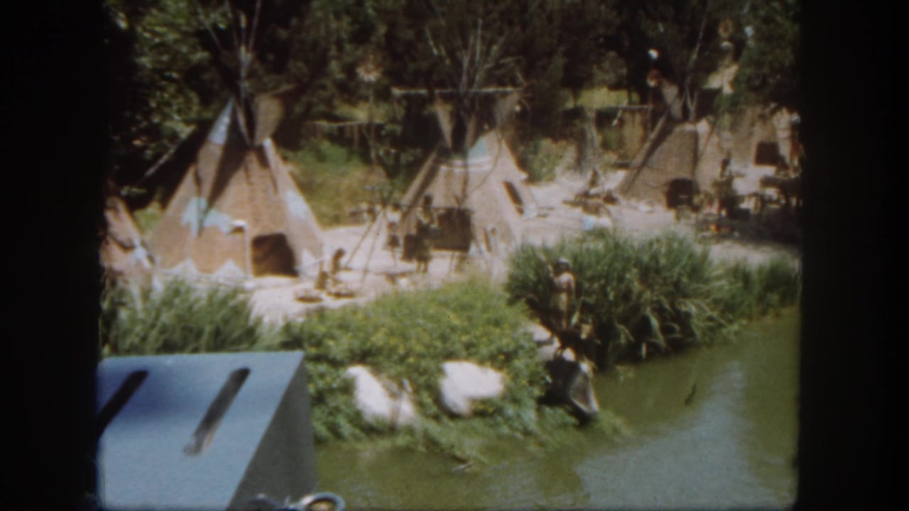 1962 Disneyland home-movie frame showing teepee-style tents, costumed figures, foliage, and a waterway in a staged village display.