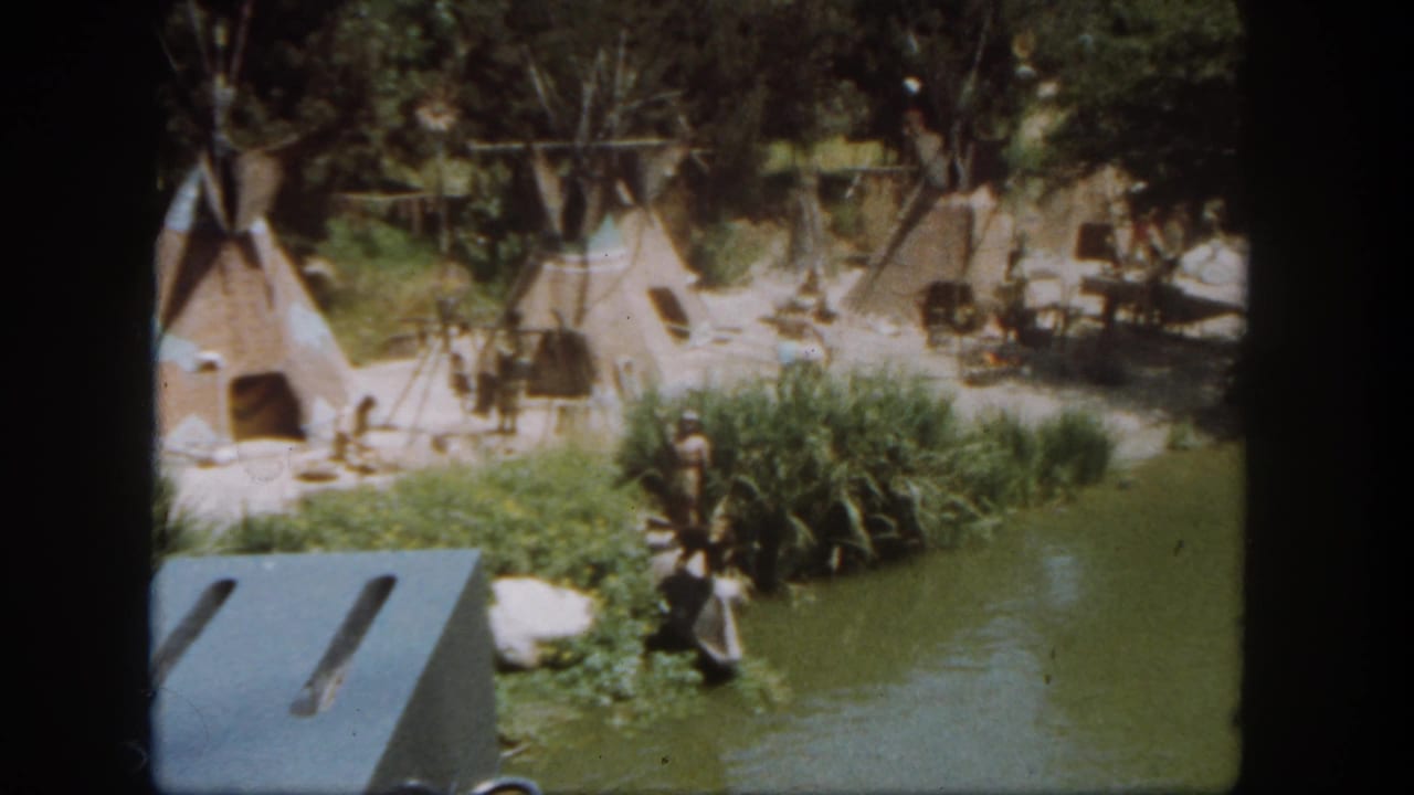 Cluster of teepee-style tents near reeds and a calm waterway, with figures on the bank.