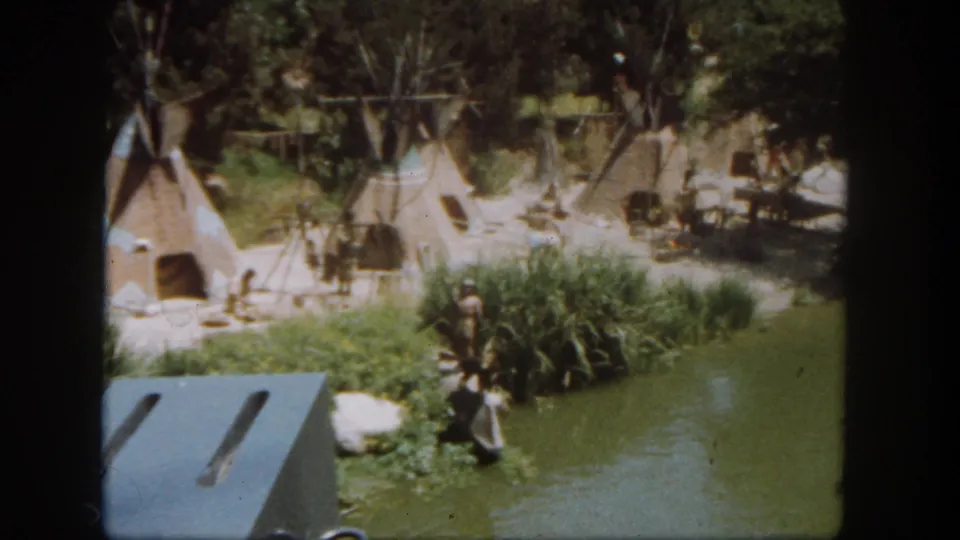 1962 Disneyland frame with teepee-style tents, figures near the riverbank, trees, reeds, and a blue-gray foreground roof edge.
