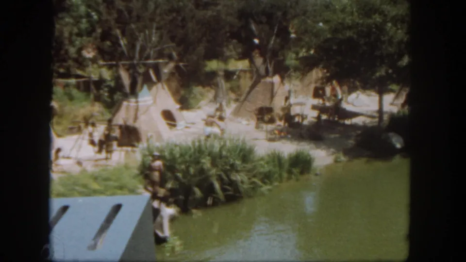 1962 Disneyland home-movie frame showing a figure by reeds and water, with staged teepee-style tents and trees behind.