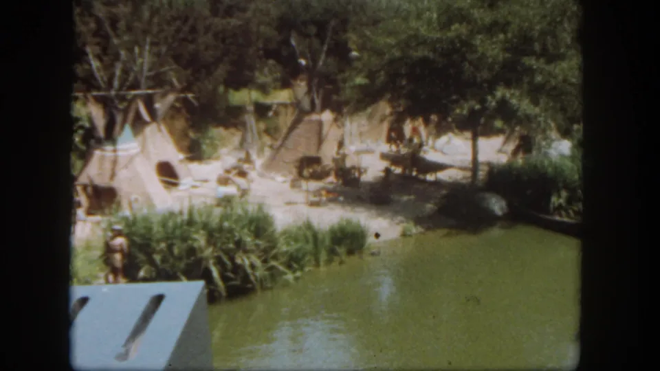 Final 1962 Disneyland frame with the waterway, reeds, trees, sandy grounds, and teepee-style tents partly framed by the pan.