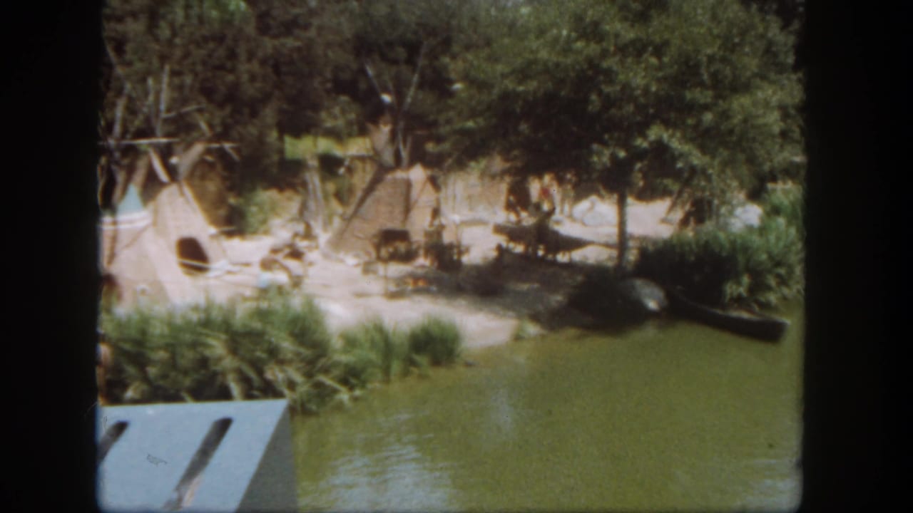 1962 Disneyland 8mm view from a boat passing Native American-style huts beside a green waterway.