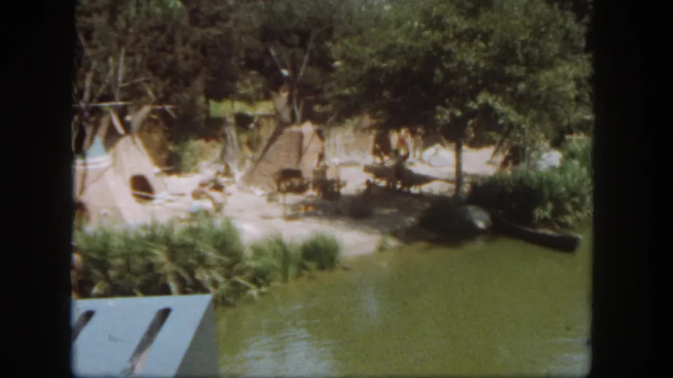 1962 Disneyland 8mm view from a boat passing Native American-style huts beside a green waterway.