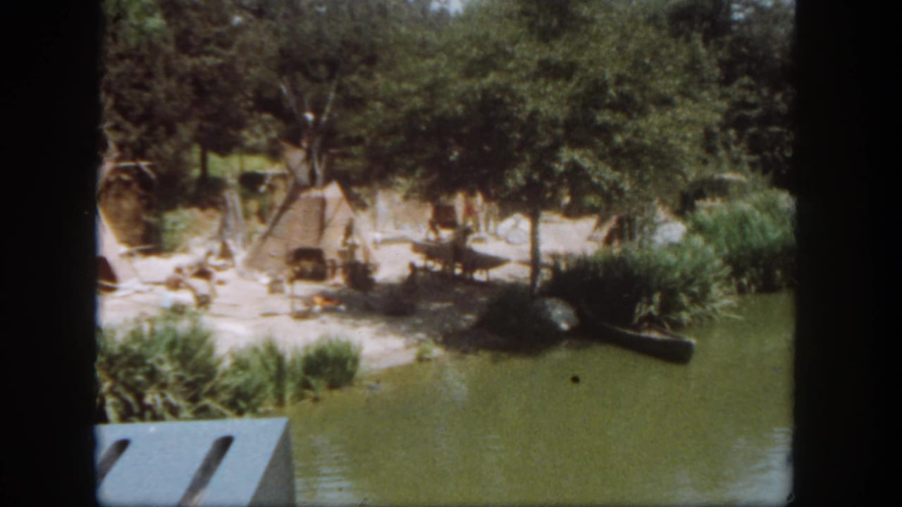 Visitor-shot 1962 Disneyland waterway scene with huts, trees, reeds, and a low shoreline structure.