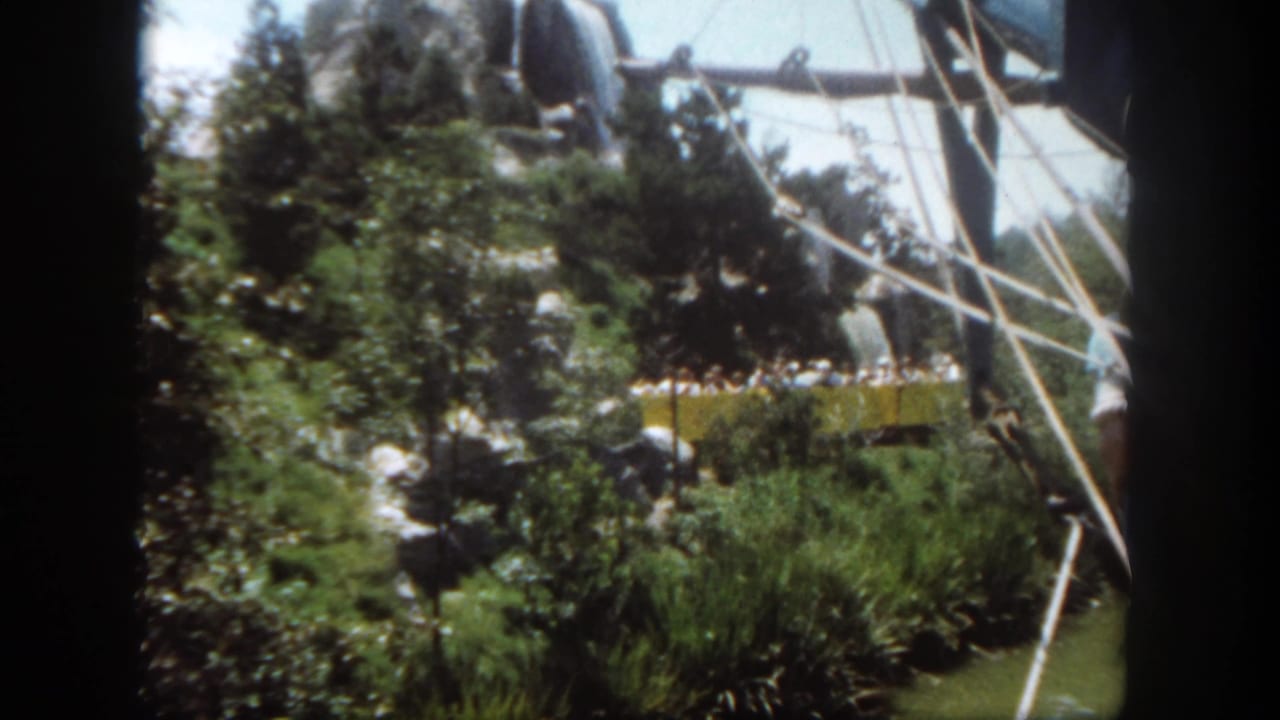 1962 Disneyland 8mm frame showing a yellow passenger tram moving through trees, rockwork, and ride framework.