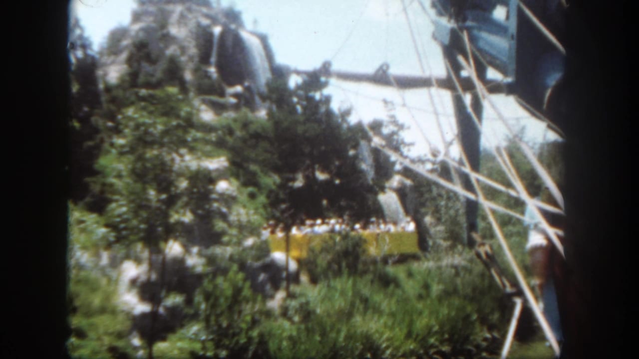 A 1962 Disneyland visitor-shot view catches a yellow tram, riders, rocks, trees, and white structural rails.