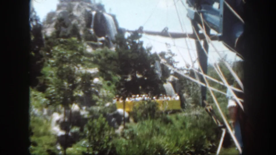 A 1962 Disneyland visitor-shot view catches a yellow tram, riders, rocks, trees, and white structural rails.