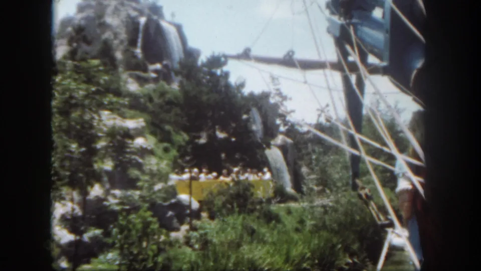 1962 Disneyland ride scenery with rock formations, green plantings, white framework, and a yellow tram partly visible below.