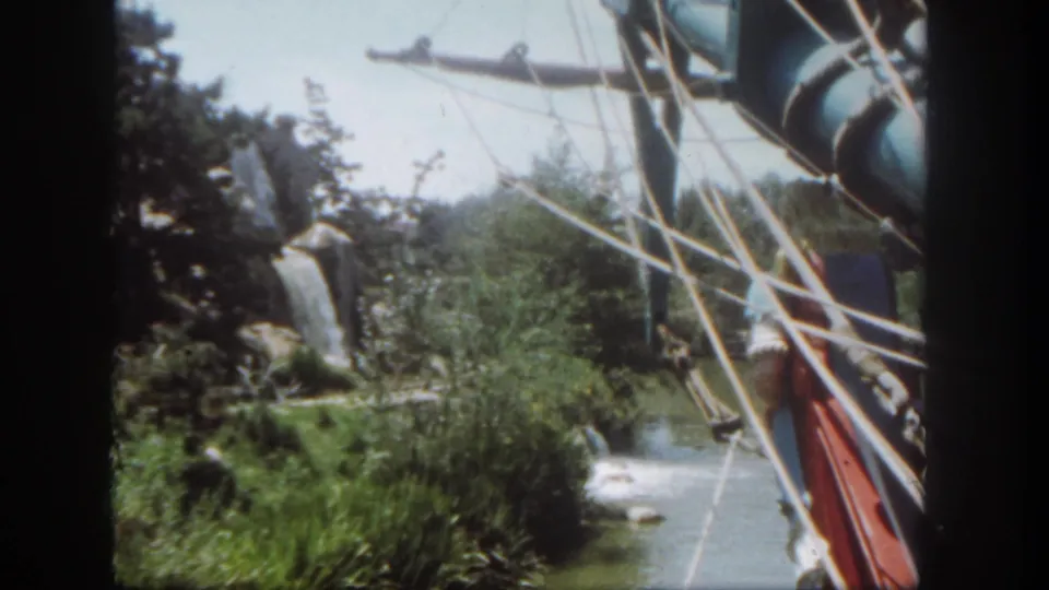 1962 Disneyland home-movie view looking past ship rigging toward dense plants, a water channel, artificial rockwork, and waterfall effects.