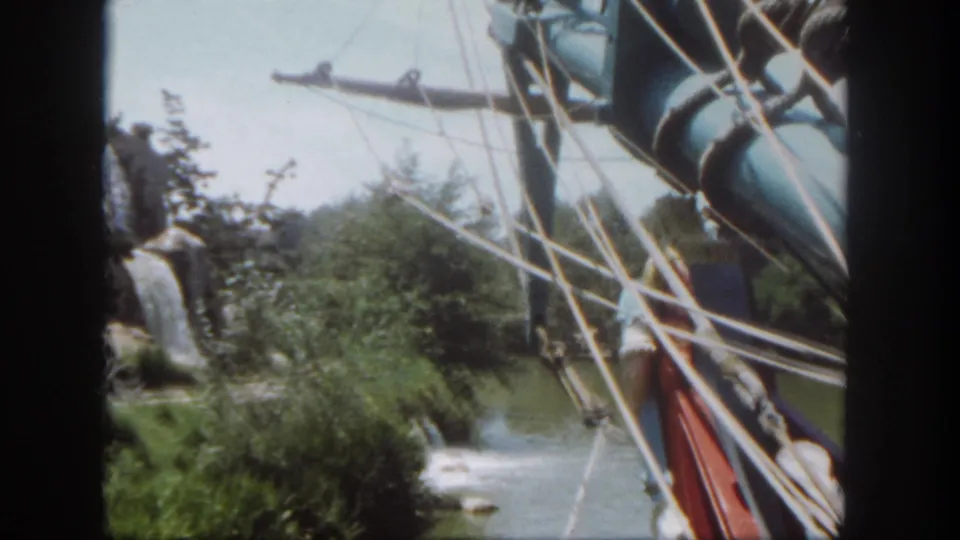 1962 Disneyland frame showing a visitor near landscaped water-ride scenery, with pirate ship rigging and a painted sail edge in the foreground.