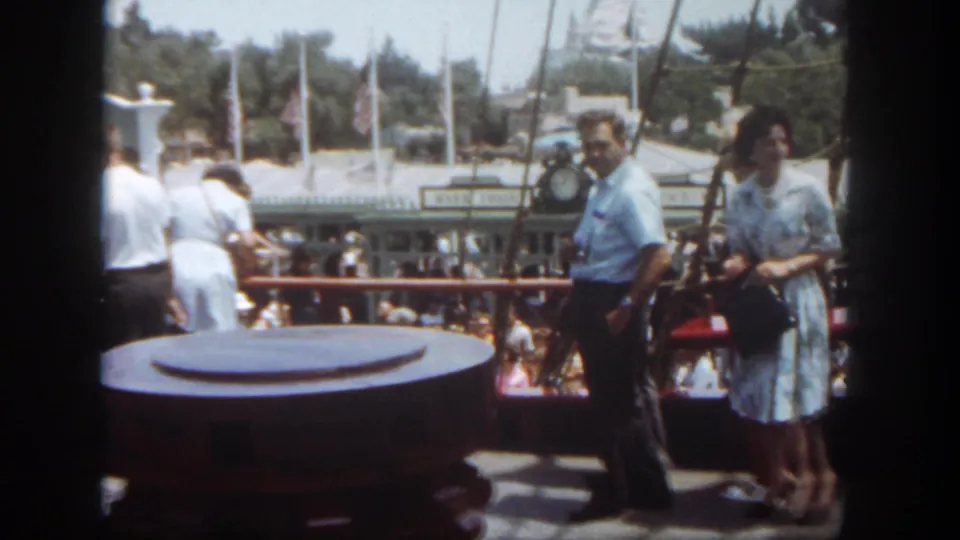 1962 Disneyland home-movie frame with guests on a riverboat deck, a large bollard in front, flags and park structures beyond.