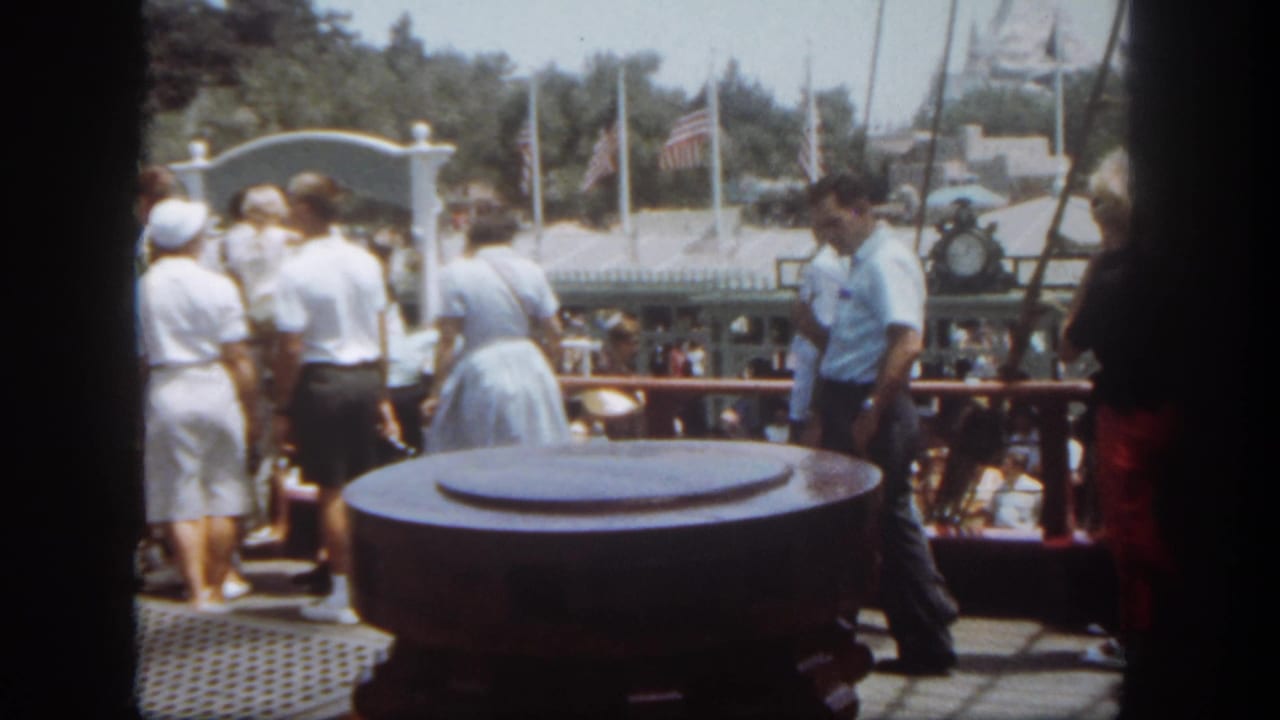 Guests in period clothing stand along a Disneyland riverboat deck, with flags, railings, and a large bollard visible.