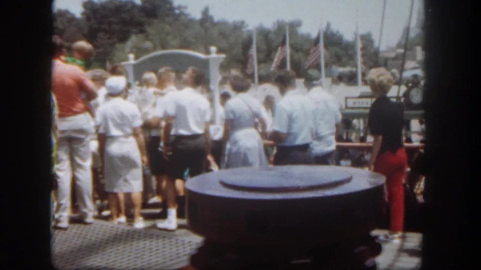 Home-movie view of Disneyland riverboat passengers gathered near the railing, with the harbor and flags visible past the deck.