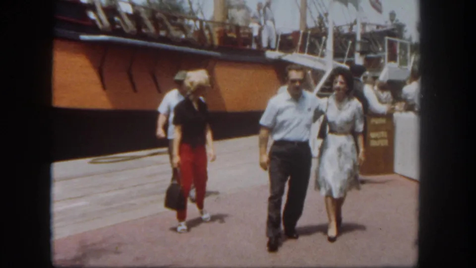 A man and woman walk along the Disneyland riverboat deck beside the wooden hull, with rigging and another visitor nearby.