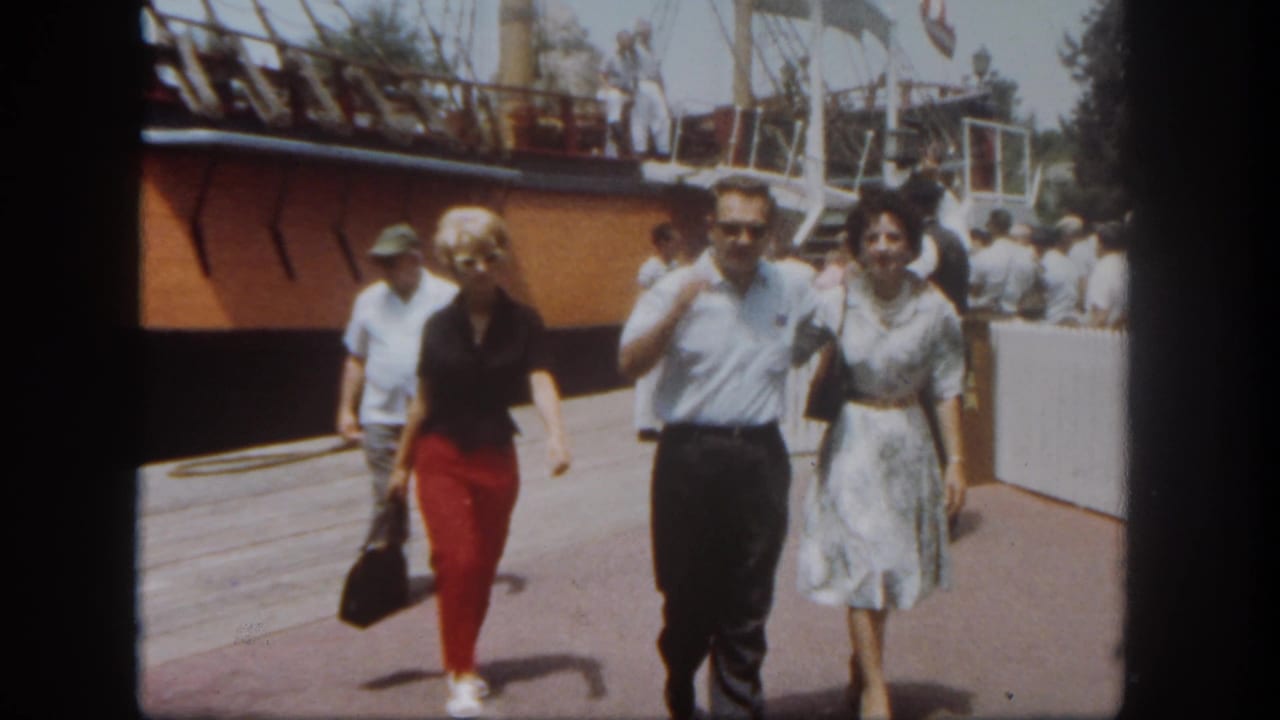 At Disneyland in 1962, adults in midcentury clothing walk along a dock area beside a large moored sailing vessel with rigging overhead.