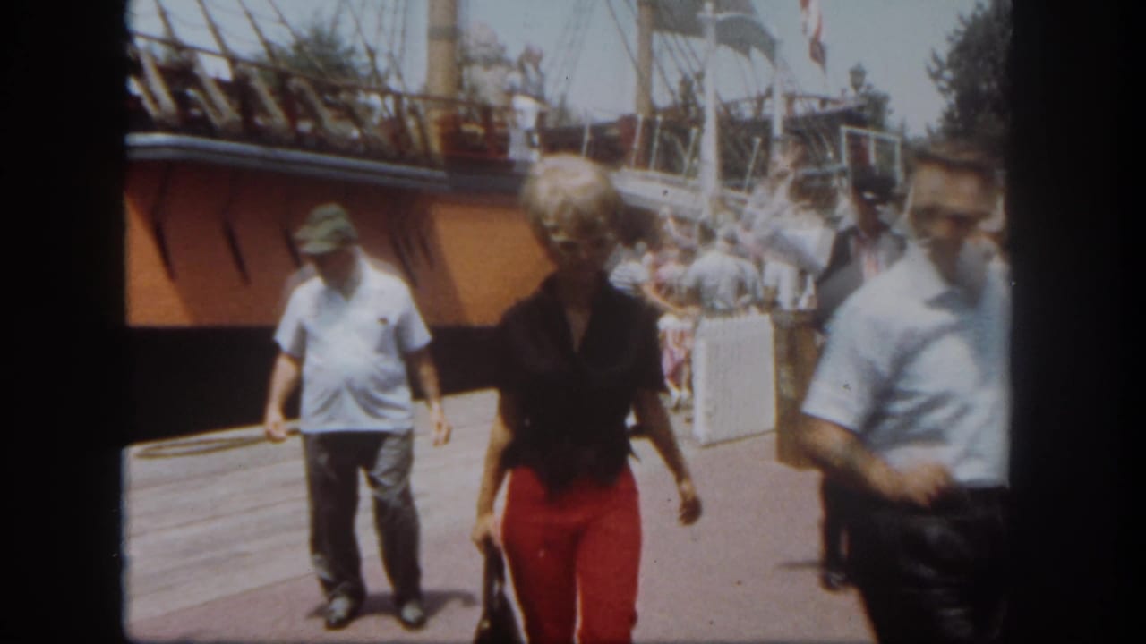 Disneyland visitors pass in front of a docked sailing vessel, with a woman in red pants, a man in a light shirt, and a ramp behind them.