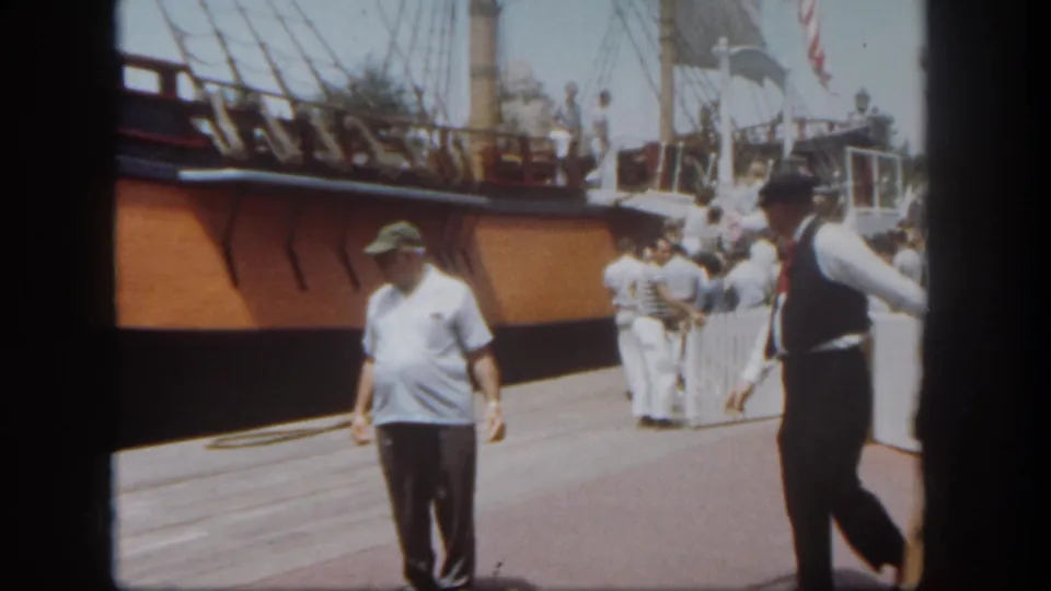 A visitor in a green hat walks past the camera at Disneyland, with a docked sailing vessel, rigging, ramp, and nearby park guests behind him.
