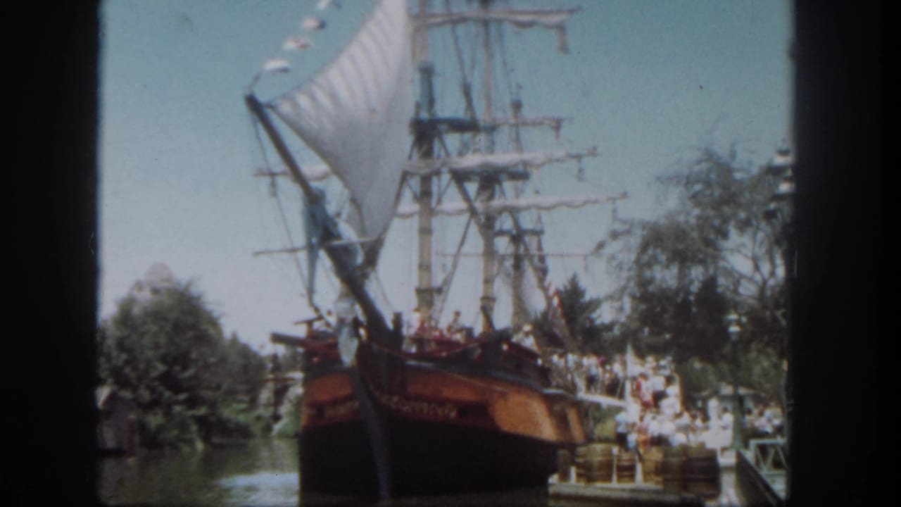 1962 Disneyland home-movie frame showing a large sailing ship attraction beside dockside guests and trees.