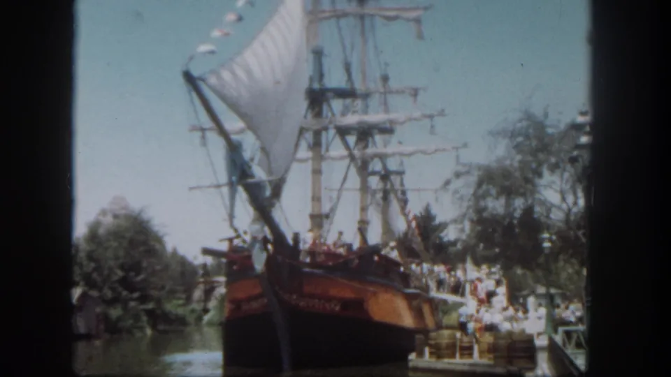 1962 Disneyland home-movie frame showing a large sailing ship attraction beside dockside guests and trees.
