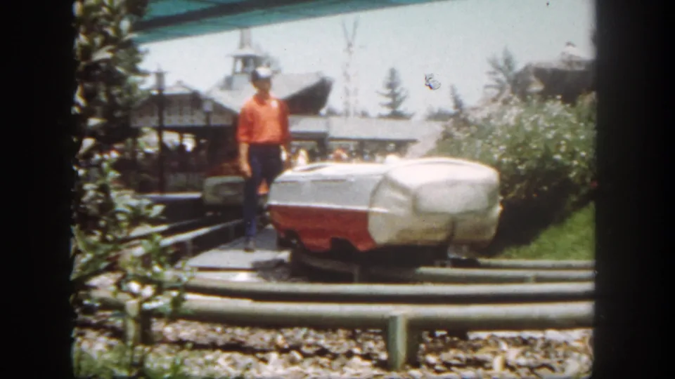1962 Disneyland home-movie frame showing an attendant beside a red-and-white boat-style ride vehicle near trees and pavilion structures.