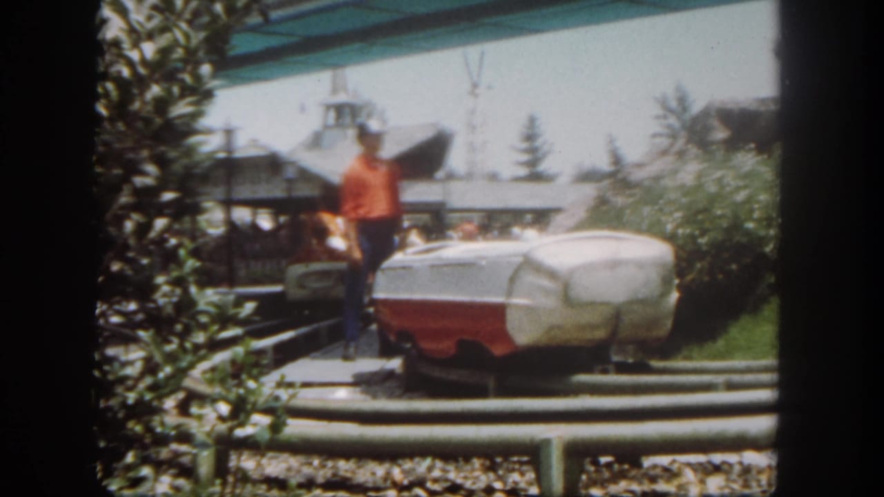 1962 Disneyland frame with an attendant in a red shirt standing by a boat-style ride vehicle on an outdoor amusement ride track.