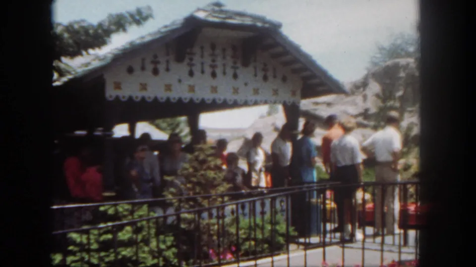 1962 Disneyland frame showing guests waiting near a decorative pavilion with scalloped trim, railing, plants, and ride area structures.