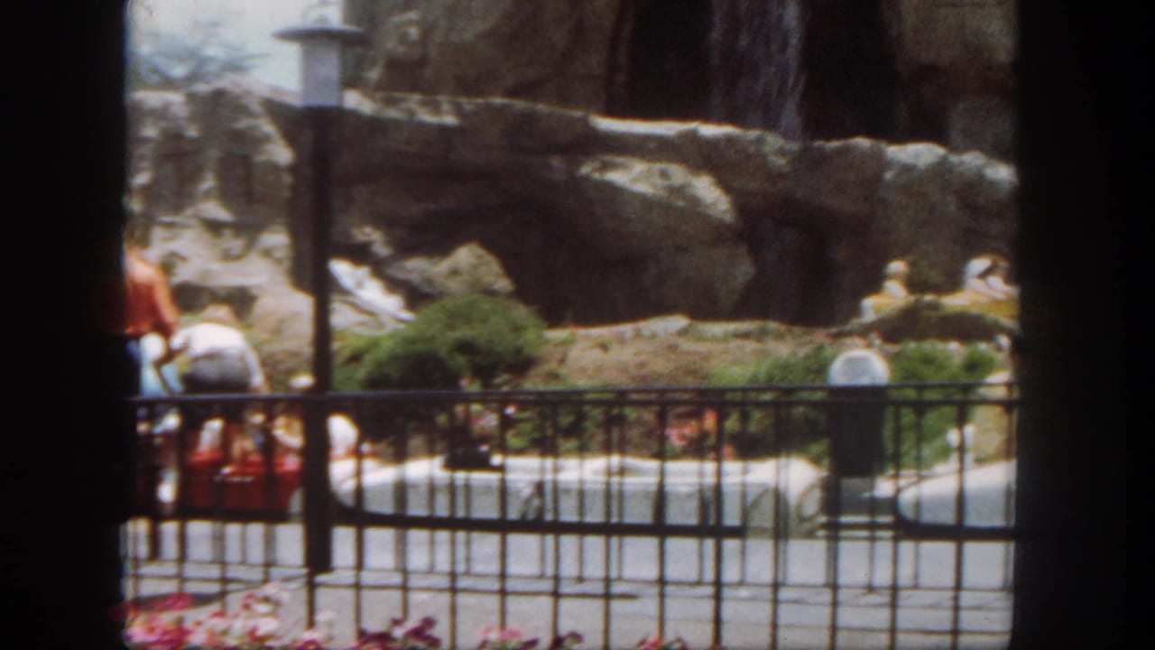 Rocky water-ride scenery behind a black metal railing with visitors and a small boat at left.