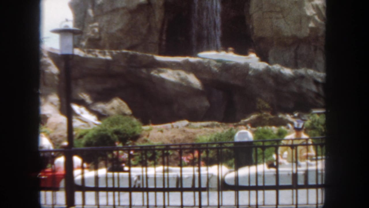 Disneyland ride scenery shows rocks, shrubs, lamps, railing, water, and a partly visible boat in the 1962 home-movie frame.