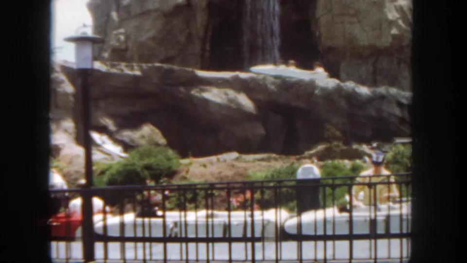 Disneyland ride scenery shows rocks, shrubs, lamps, railing, water, and a partly visible boat in the 1962 home-movie frame.