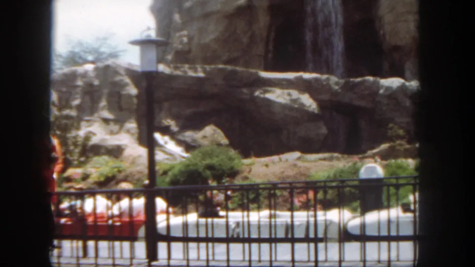 1962 Disneyland home-movie frame looking past a black railing toward rockwork, shrubs, water, and a partly visible ride boat.
