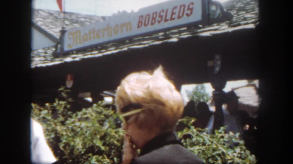 Disneyland frame with the Matterhorn Bobsleds sign, shrubs, roofline, and a blonde visitor in sunglasses near the foreground.