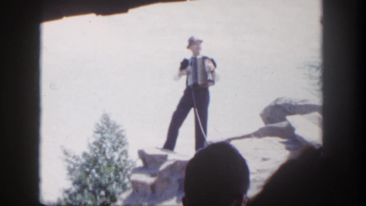 1962 Disneyland frame showing a suited man in a hat playing accordion on a rocky outcrop before silhouetted spectators.