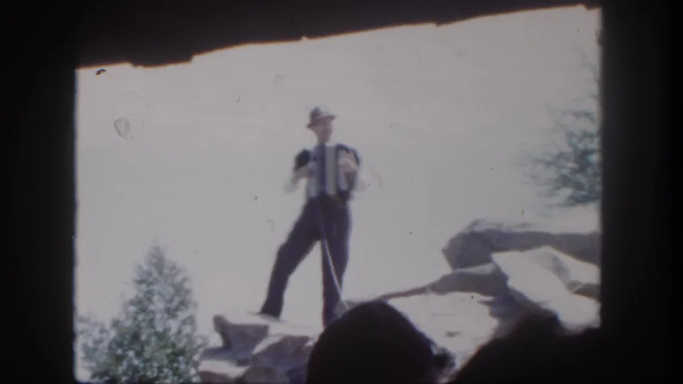 1962 Disneyland home-movie frame of a hat-wearing accordionist standing on rocks, with soft focus and dark frame edges.
