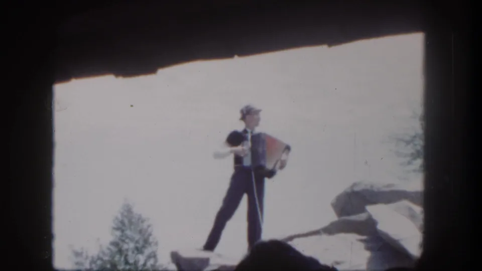 1962 Disneyland frame of a suited accordion player on a rocky set, seen above dark audience silhouettes in bright daylight.