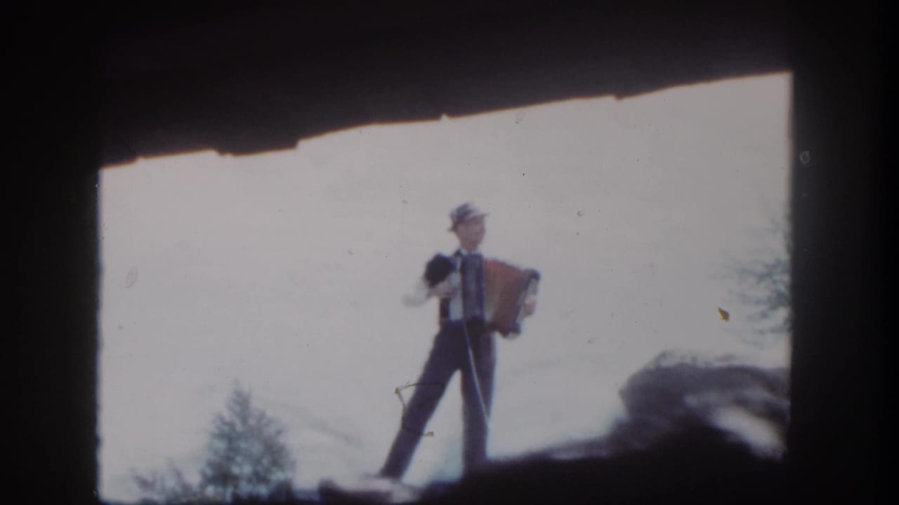 Man in a dark suit and hat plays an accordion on a rocky ledge under a bright sky.