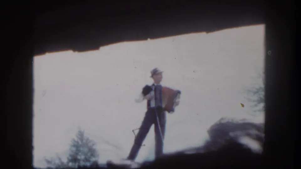 At Disneyland in 1962, a suited accordion player stands on a rocky ledge with trees low in the background.