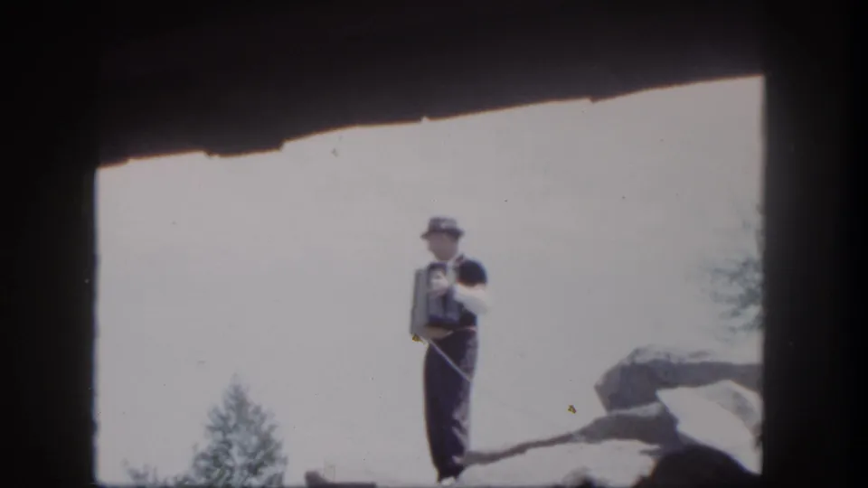 The 1962 Disneyland home-movie frame shows the accordion performer standing upright beside a rocky set piece.