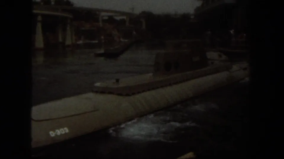Darker 8mm frame showing a submarine marked D-303 on the water, with ride structures and shoreline visible beyond.